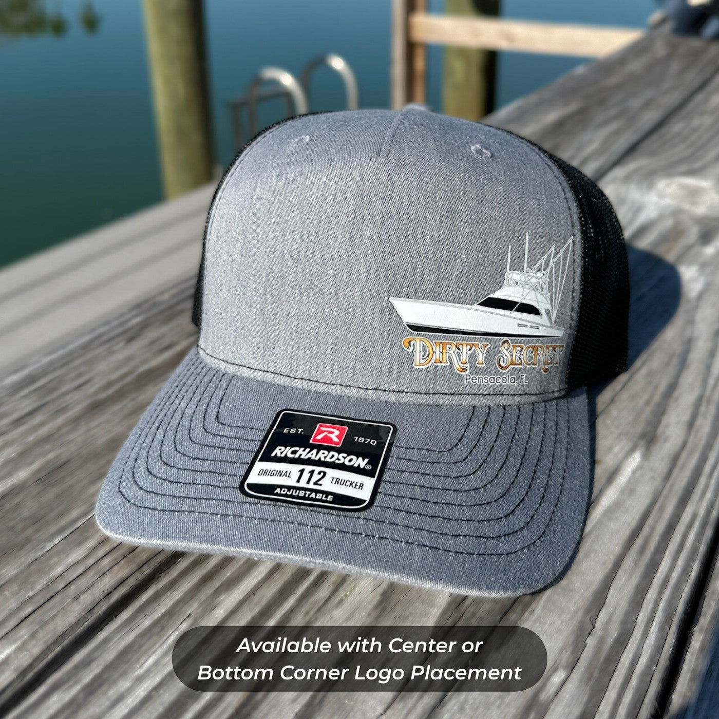 Gray trucker hat with boat graphic and Richardson logo on a wooden surface by water.