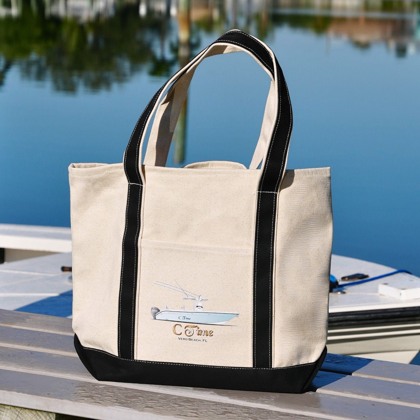 Beige tote bag with custom printed boat, black handles and trim on a dock by water