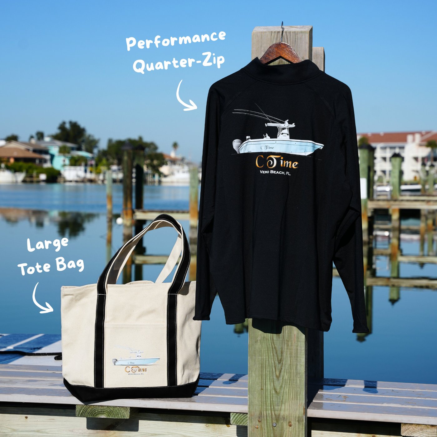 Black performance quarter-zip shirt and large tote bag with custom boat branding on a dock with water and buildings in the background.